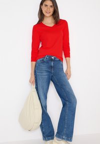 Maglietta rossa a maniche lunghe con scollo a V abbinata a jeans blu a vita alta e svasati. Il modello tiene una borsa a tracolla beige e indossa scarpe di colore chiaro.