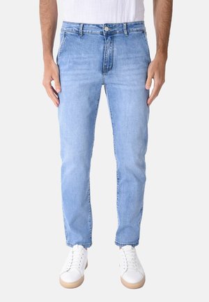 Uomo con jeans slim fit color azzurro chiaro e sneakers bianche, in piedi davanti a uno sfondo chiaro e uniforme.