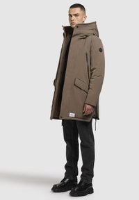 Parka izolată de culoare marron, cu guler înalt, buzunare cu fermoar și un exterior texturat. Purtați peste un top negru și asociați cu blugi negri și cizme.