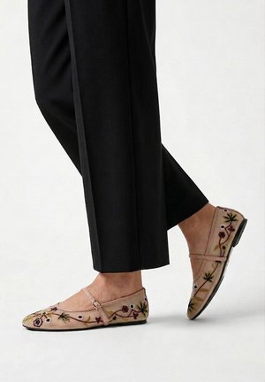 Pies con bailarinas rosa claro con bordado floral y una tira, combinados con pantalones negros hasta el tobillo sobre un fondo liso.