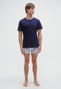 T-shirt de algodão azul-marinho com decote redondo e mangas curtas, combinada com cuecas boxer justas cinza claro. Modelo em pé, descalço.