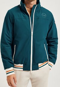 Teal vindjacka med vit dragkedja, kontrasterande randiga ärmar i orange och vitt, samt en logotyp på bröstet. Slät textur.