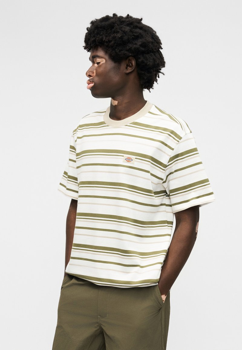 Junger Mann mit lockigem Haar, der in gestreiftem weiß-olivgrünem T-Shirt und olivgrüner Hose steht, Hände in den Taschen, nach links blickend vor einfarbigem Hintergrund.