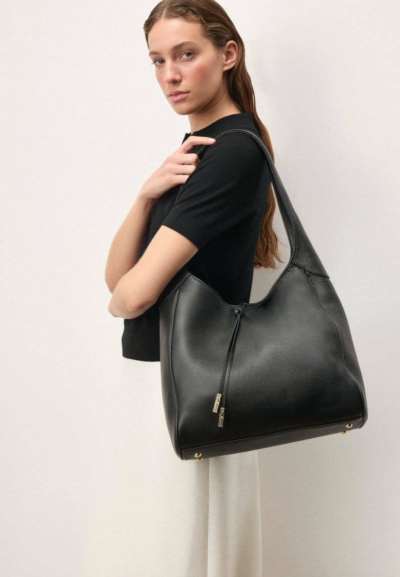 Kobieta ubrana w czarną bluzkę z krótkim rękawem i jasną spódnicę trzyma dużą czarną skórzaną torbę na ramię ze złotymi akcentami.