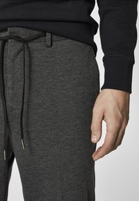 Szare sportowe spodnie dresowe z sznurkiem w pasie, zestawione z ciemnym swetrem. Widoczna faktura materiału oraz dłoń spoczywająca na pasku w talii.