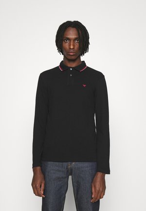 Svart långärmad polotröja med krage, röd och vit accent på kragen, och en liten röd logotyp på bröstet. Bärs med blå jeans.