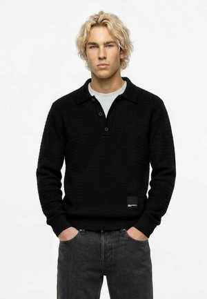Junger Mann mit blondem Haar, der einen schwarzen strukturierten Pullover über einem weißen Hemd und dunklen Jeans trägt, steht mit den Händen in den Taschen vor weißem Hintergrund.