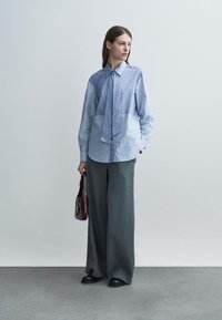 Nainen, jolla on sinivalkoraidallinen paita edestä solmittuna, harmaat leveälahkeiset housut, mustat kengät ja joka pitää tummaa ruskeaa käsilaukkua, seisoo yksiväristä taustaa vasten.