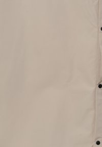 Beżowa kurtka wierzchnia wykonana z materiału syntetycznego, o gładkiej teksturze, zapinana na zatrzaski, bez widocznych wzorów ani akcentów.