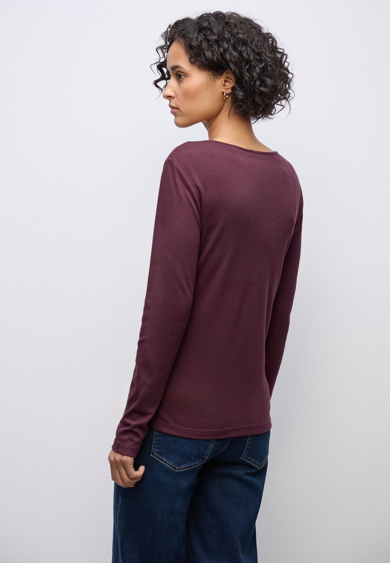 Weinrotes Langarmoberteil mit rundem Halsausschnitt und taillierten Ärmeln, kombiniert mit dunkelblauen weitgeschnittenen Jeans, von hinten gezeigt.