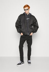 Giubbotto bomber nero con zip, indossato sopra un felpa scura con cappuccio. Jeans neri e scarpe Vans nere completano il look. Modello con occhiali da sole.