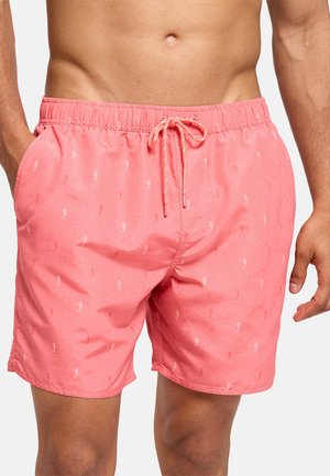 Hombre con pantalones cortos de baño de color coral rosa con diseño de caballitos de mar y cintura con cordón, manos parcialmente visibles, torso mostrado desde el ombligo hasta la mitad del muslo.