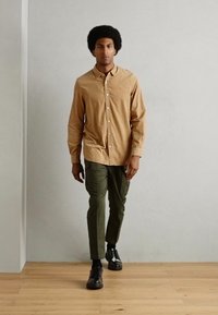 Camisa de botón marrón claro con mangas largas y un logo en el pecho, combinada con pantalones tapered verde oliva y zapatos negros sobre un suelo de madera.