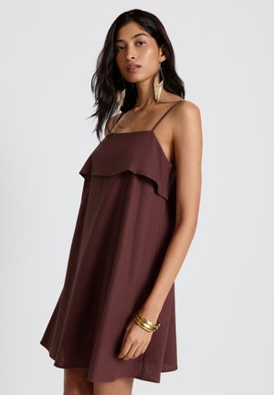 Donna che indossa un vestito marrone senza maniche con sottili spalline, braccialetti d'oro e lunghi orecchini d'oro, in piedi davanti a uno sfondo chiaro e semplice.