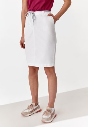 Vrouw draagt een witte rok tot op de knie met een taillekoord, gecombineerd met beige en witte sneakers en een donkerroze top.
