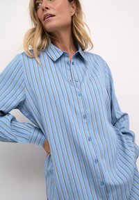 Light blue button-up shirt with vertical white and gold stripes. Features a collar, long sleeves, and a relaxed fit.