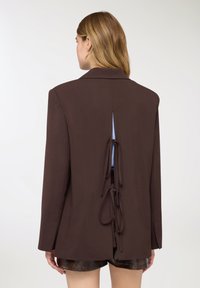 Blazer marrone con colletto a risvolto, schiena scoperta con inserto azzurro e dettagli annodati. Tessuto liscio, vestibilità aderente, maniche lunghe.