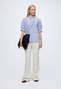 Femme portant un chemisier bleu clair à manches bouffantes, un pantalon blanc à jambes larges, des sandales noires, tenant une pochette noire à franges.