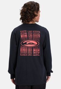 Schwarzes langärmliges T-Shirt mit einem rosanen Grafikdesign auf der Rückseite, das den Text "Schmeckt nach Himmel" in einer wellenförmigen, verzerrten Schriftart zeigt.