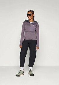 Vijolična zip-up jakna z teksturirano tkanino, z dvema sprednjima žepoma, v kombinaciji s črnimi hlačami in zelenimi pohodniškimi čevlji, na nevtralni podlagi.