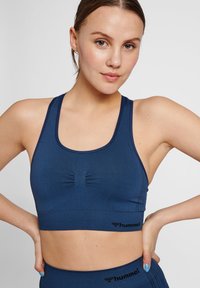 Marinblå sport-bh med en figurnära design, som har en samlad detalj i mitten och en logotyp-accent. Slät, elastisk tyg.