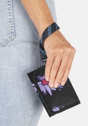 Hand hält schwarze Stoffgeldbörse mit lila Blumendesign und schwarzem Riemen vor hellblauem Jeansstoffhintergrund.