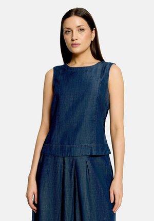 Woman with straight dark hair wearing a sleeveless dark blue denim top and matching pleated skirt against a plain light background.