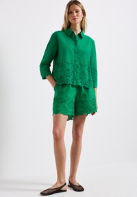 Zelená lněná košile se zubatým lemem, s výřezovými vzory a zapínáním na knoflíky, kombinovaná s odpovídajícími zubatými šortkami. Černé síťované klouzavé boty.