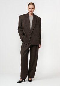 Egy nő oversize sötétbarna öltönyben, széles vállakkal, hozzávaló bőszárú nadrágban, szürke felsőben, fekete magassarkúban és arany fülbevalókban.