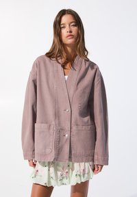 Overmaat roze katoenen jas met een V-hals, twee voorzakken en knoopsluiting. Draagt over een gelaagde bloemenrok in wit en groen.