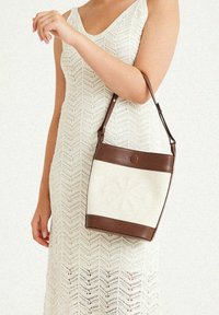 Weißer Canvas-Handtasche mit braunem Lederbesatz und -riemen, mit einem blumenartigen geprägten Design auf der Vorderseite.