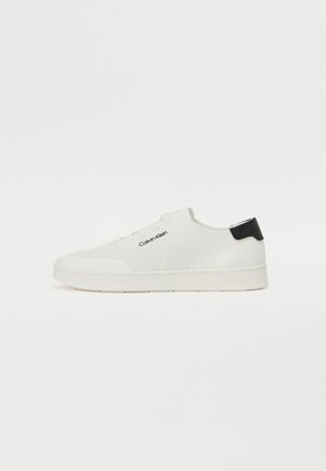 Bílá nízká teniska s pletenou texturou, černou patní záložkou a logem „Calvin Klein“ na boku, zobrazena na bílém pozadí.
