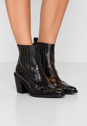 Bottines en cuir motif serpent noir et marron, bouts pointus et talons blocs, portées sur jambes nues.