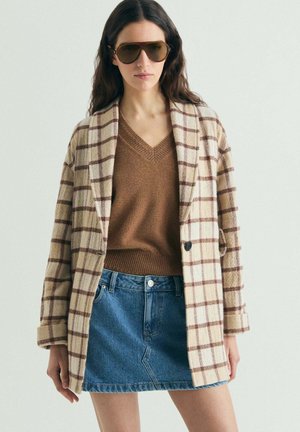 Mujer con abrigo grande de cuadros beige, suéter marrón de cuello en V, minifalda de mezclilla y grandes gafas de sol marrones sobre un fondo liso.