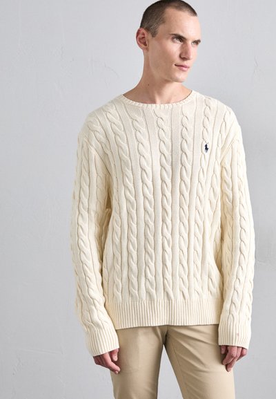 Cream cable-knit sweater with ribbed cuffs and hem, featuring a small embroidered logo on the left chest. Relaxed, oversized fit.
