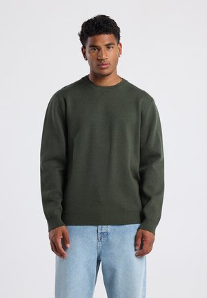 Pull à col rond vert foncé en tissu doux et texturé, avec des poignets et un ourlet côtelés, associé à un jean bleu clair.