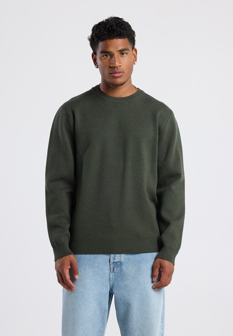 Dark green crew-neck sweater in a soft, textured fabric with ribbed cuffs and hem, paired with light blue jeans.