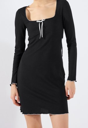 Czarna żebrowana sukienka z długim rękawem, kwadratowym dekoltem, białym przeszyciem na krawędziach oraz małą białą kokardką na środku z przodu.