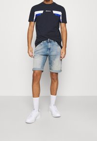 Mężczyzna w granatowej koszulce z grafiką, jasnoniebieskich spodni denimowych z podwiniętymi nogawkami, białych skarpetkach i białych trampkach, stoi na tle jednolitego tła.