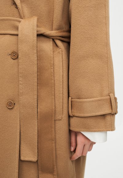 Primo piano di un cappotto in lana color cammello con cintura, dettagli con bottoni, tasca laterale e cinturino al polsino su una manica di camicia bianca.