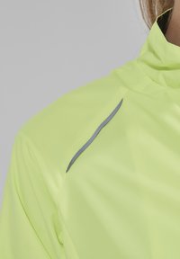 Světlá žlutá sportovní bunda s vysokým límcem a šedým reflexním akcentem na rameni. Hladký, lehký materiál s texturovaným povrchem.