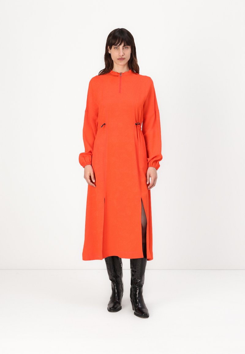 Lange oranje jurk met hoge kraag, ritssluiting en wijd uitlopende mouwen. Voorzien van zijsplitten en koorddetails. Graag gedragen met zwarte kniehoge laarzen.