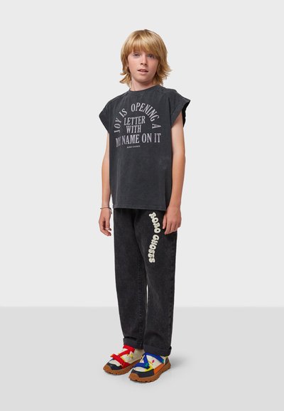 Camiseta sin mangas negra con texto impreso, combinada con pantalones anchos negros que presentan un logo blanco; zapatillas coloridas con patrones mezclados y correas.