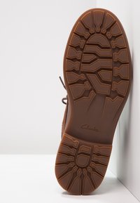 Bota de cuero marrón con una suela de goma Texturizada. La suela presenta un patrón de banda de rodadura detallado, un logo "Clarks" grabado y una punta redondeada.