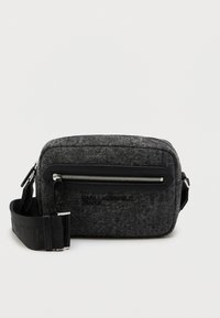 Bolsa transversal cinza de feltro com um bolso frontal com fecho zip, logótipo preto e correia ajustável. Possui uma forma retangular estruturada e hardware metálico suave.