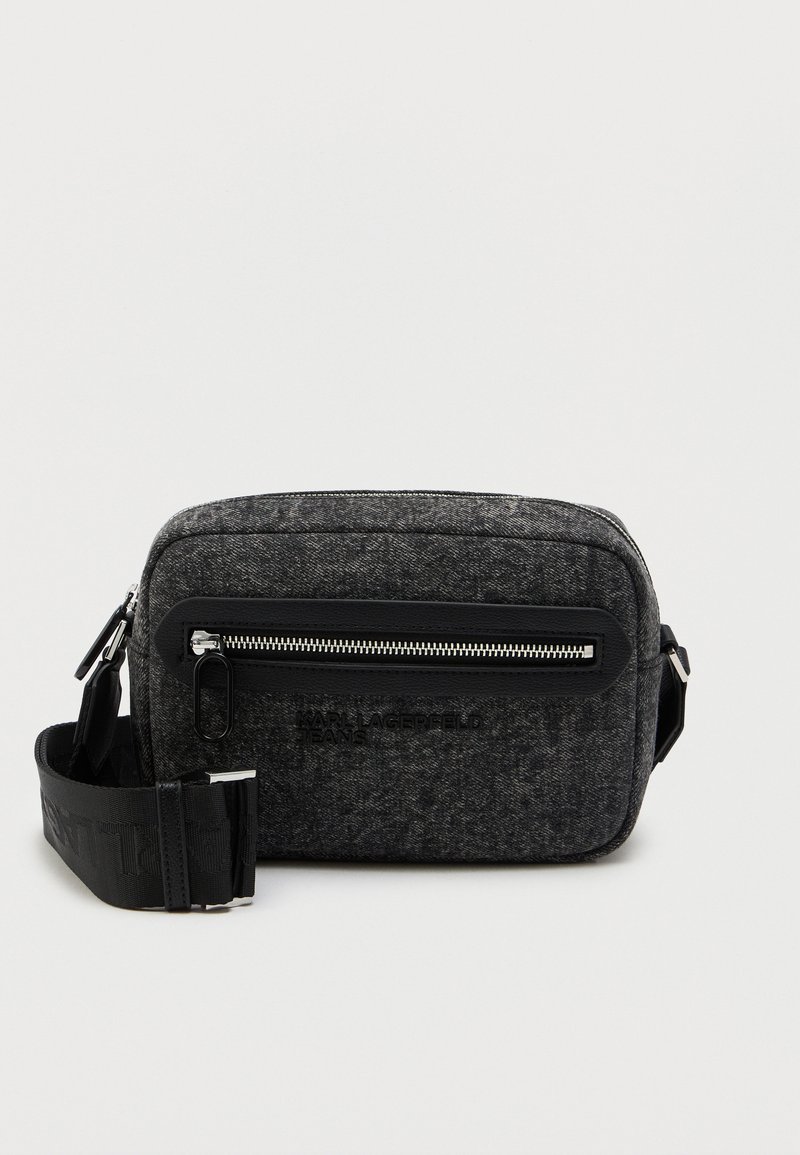 Bolsa transversal cinza de feltro com um bolso frontal com fecho zip, logótipo preto e correia ajustável. Possui uma forma retangular estruturada e hardware metálico suave.