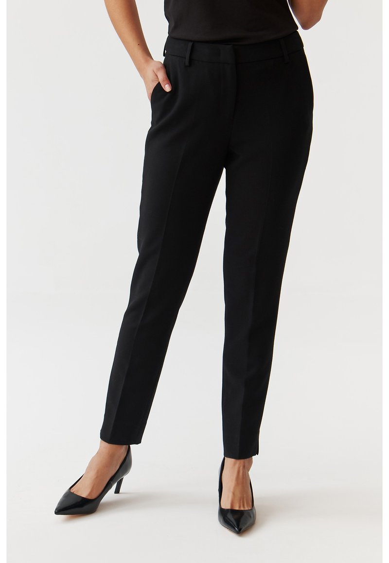 Pantaloni neri aderenti in un tessuto liscio, con tasche laterali e un design sartoriale, abbinati a décolleté nere a punta.