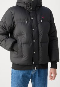 Schwarze Daunenjacke mit Kapuze, ausgestattet mit silbernen Druckknöpfen, Fronttaschen und gerippten Bündchen. Mittellang mit einer strukturierten Oberfläche.