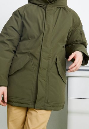 Persoon draagt een olijfgroene gewatteerde jas met grote voorzakken en een beige broek, leunend op een wit oppervlak.