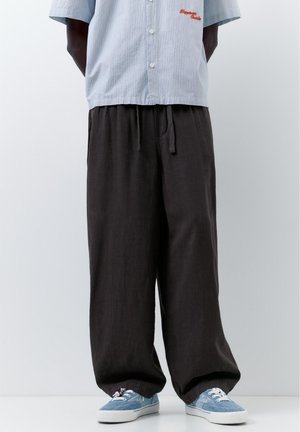 Person trägt lockere schwarze Hose, hellblau gestreiftes Kurzarmhemd und hellblaue Sneakers vor schlicht weißem Hintergrund.